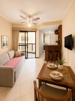 A living room with a sofa, a dining table set for two, a kitchen area, and a balcony with a sliding door. - Fully equipped apartment in Campinas with pool, gym, and garage. (Campinas)