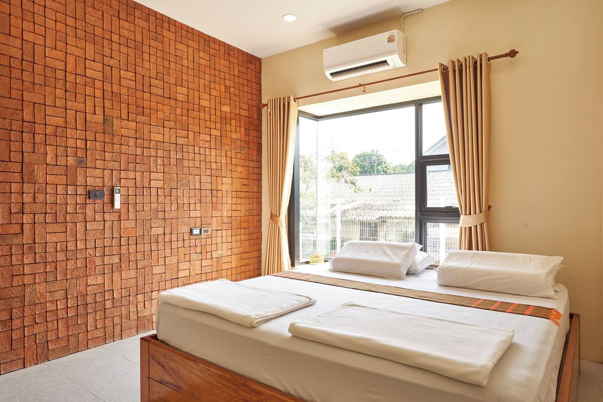 A modern bedroom with a brick wall, a bed with white linens, an air conditioning unit, and a window with curtains.