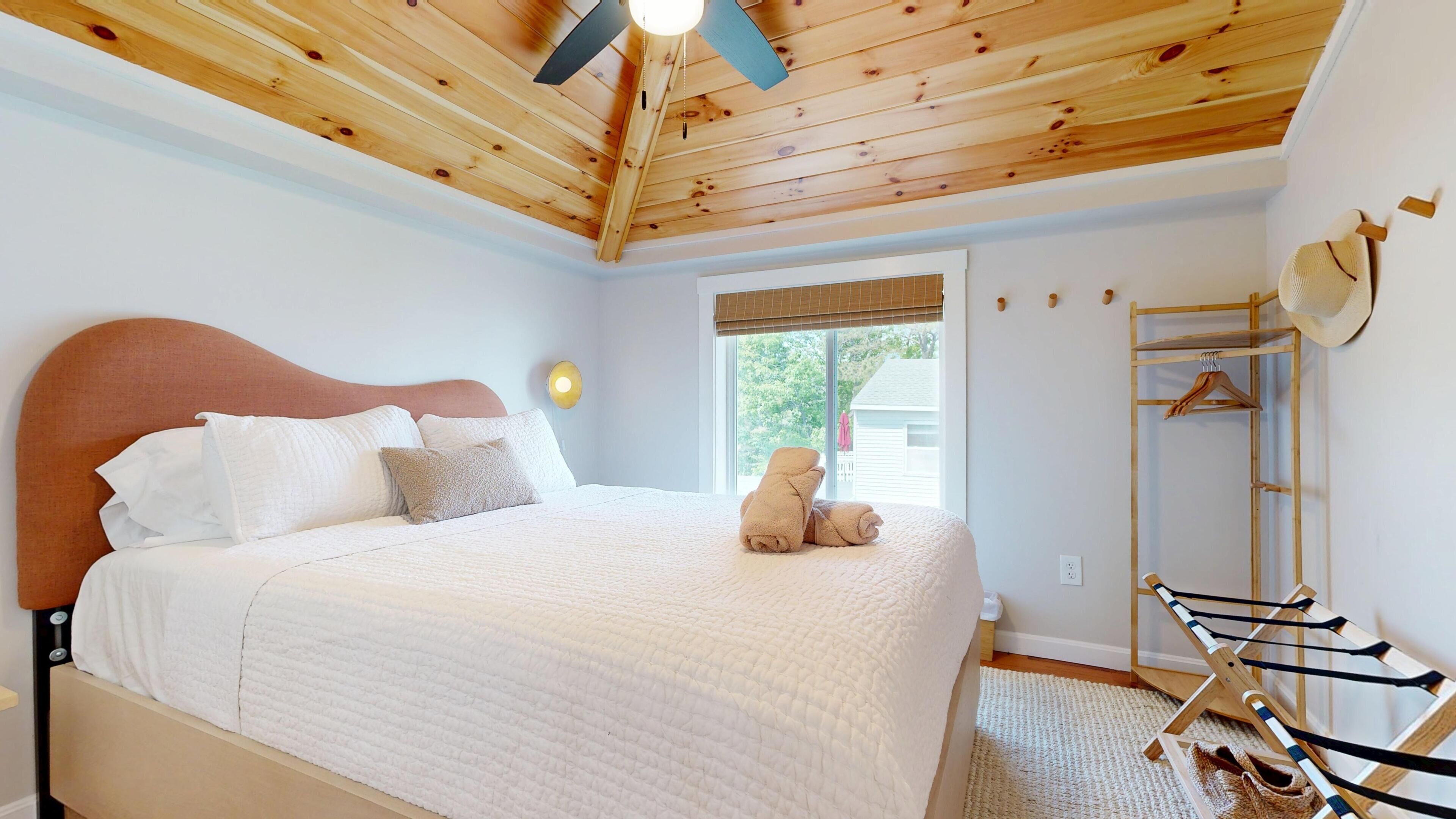 Une chambre avec un plafond en bois, un lit avec une literie blanche, une fenêtre avec des stores et un étendoir à linge en bois.