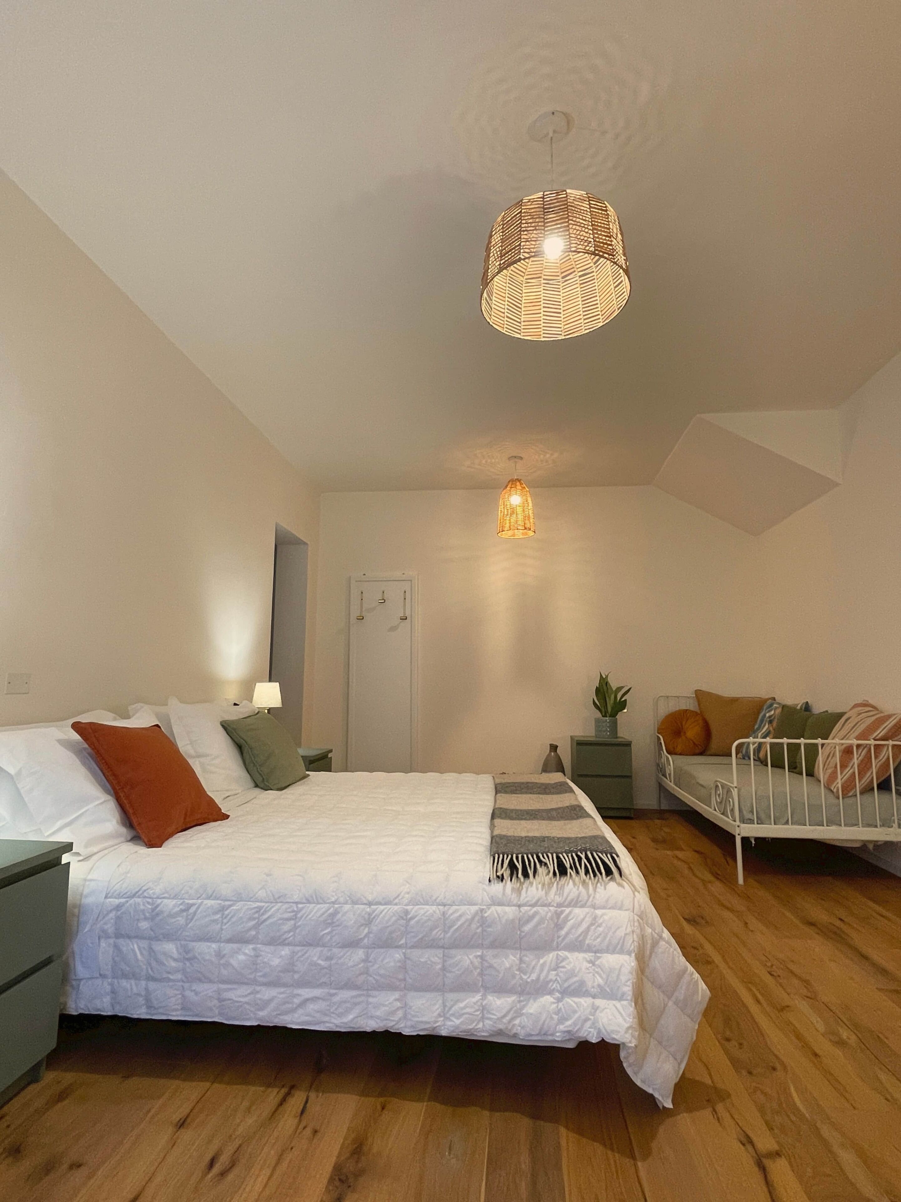 Een slaapkamer met een bed, nachtkastjes, een hanglamp, een deur en een kleine plant.