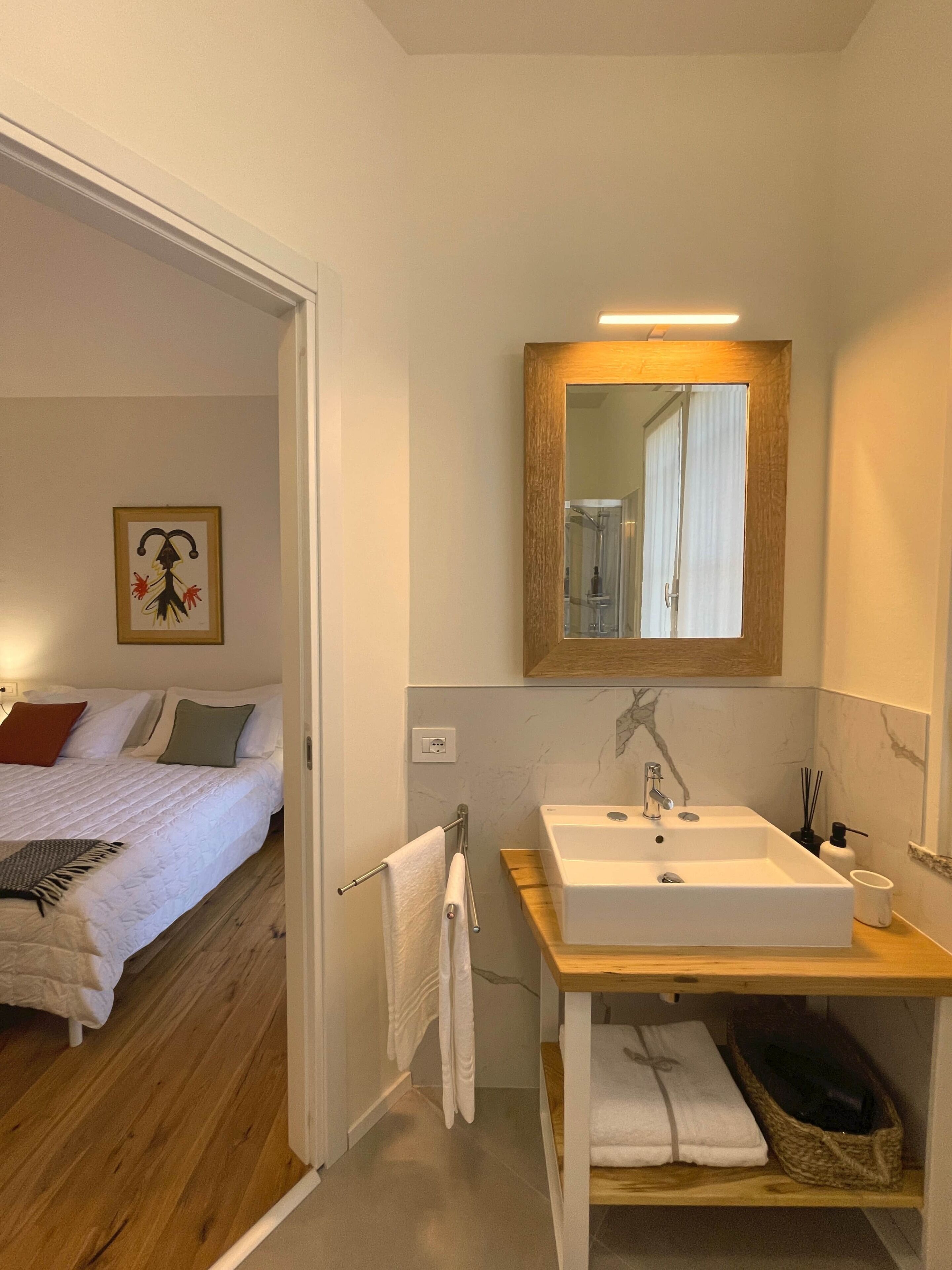 A hotel room with a bed, a wooden floor, a white sink, a mirror, and a small wooden shelf with towels.