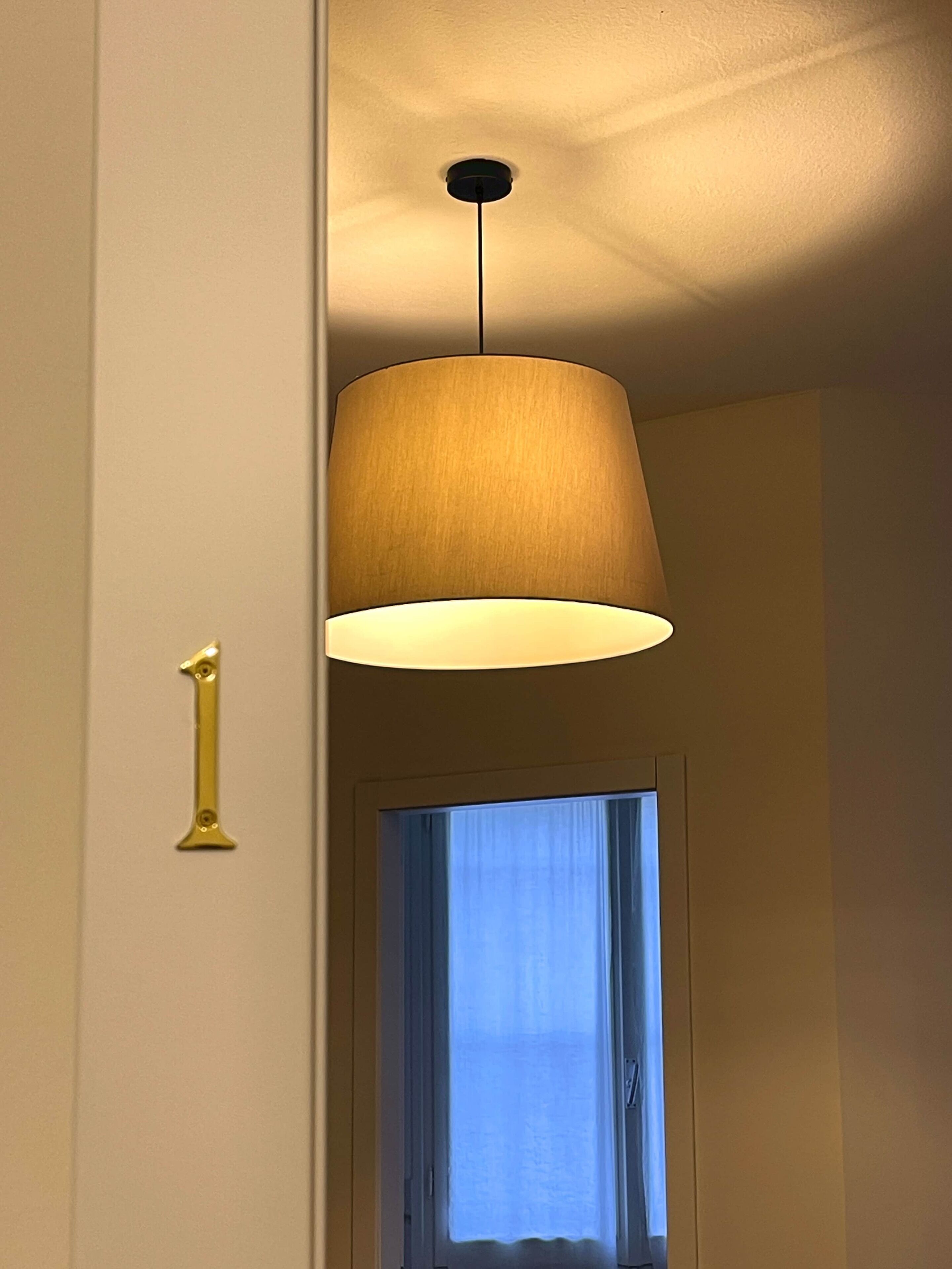 Een gang met een deur waarop een messing nummer '1' staat, een hangende lamp en een raam met gordijnen.
