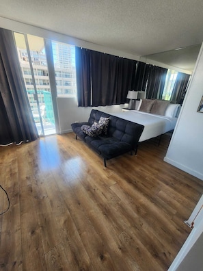 A modern hotel room with a wooden floor, a black sofa, a bed, and a large window with curtains. - 9th Floor Studio in the Middle of Waikiki (Honolulu)