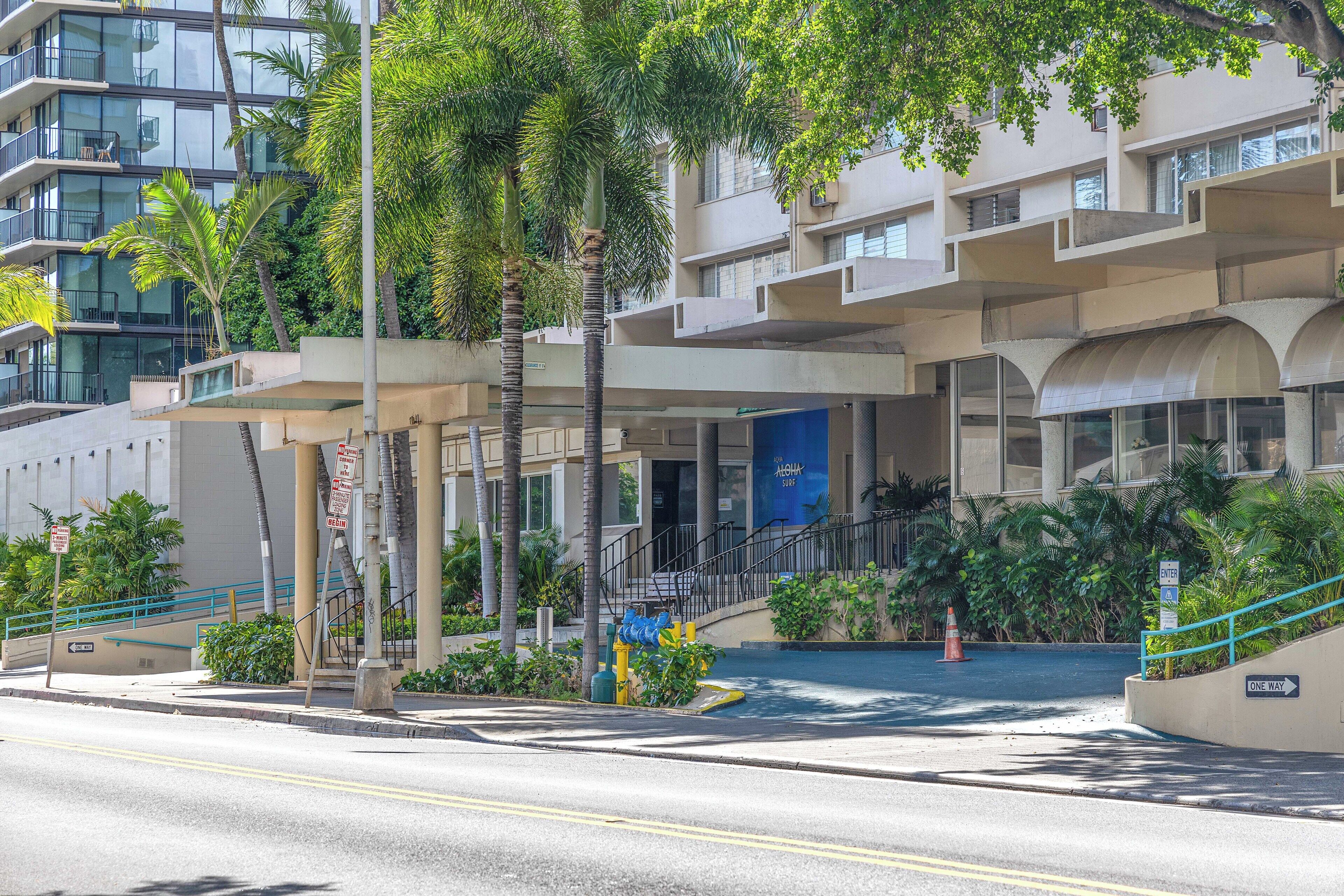 A multi-story hotel with a modern architectural design, featuring balconies, a covered entrance, and a landscaped area with palm trees.