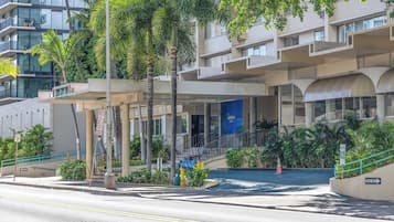 A multi-story hotel with a modern architectural design, featuring balconies, a covered entrance, and a landscaped area with palm trees.