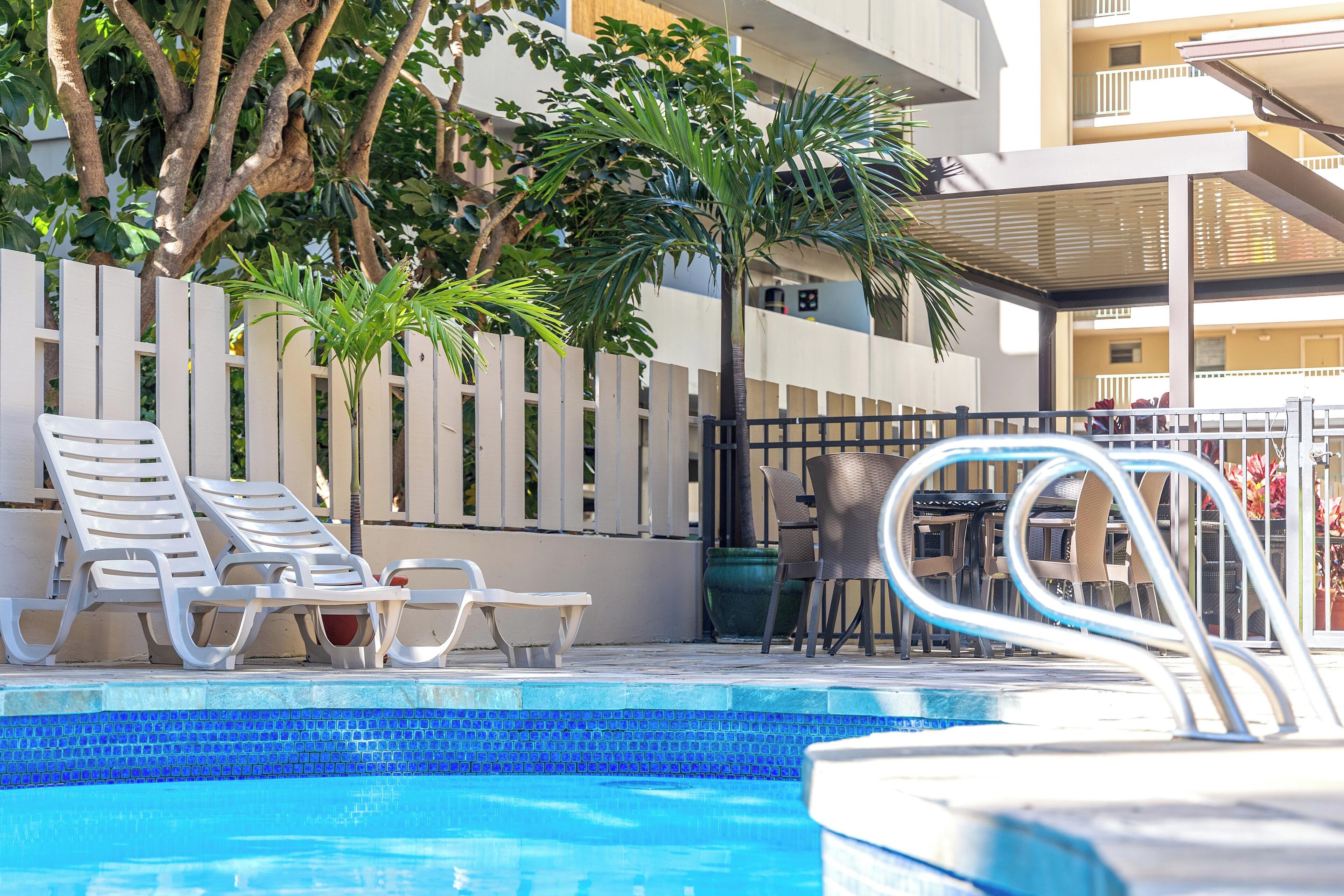 A swimming pool area with lounge chairs and a table, surrounded by a white fence and greenery.