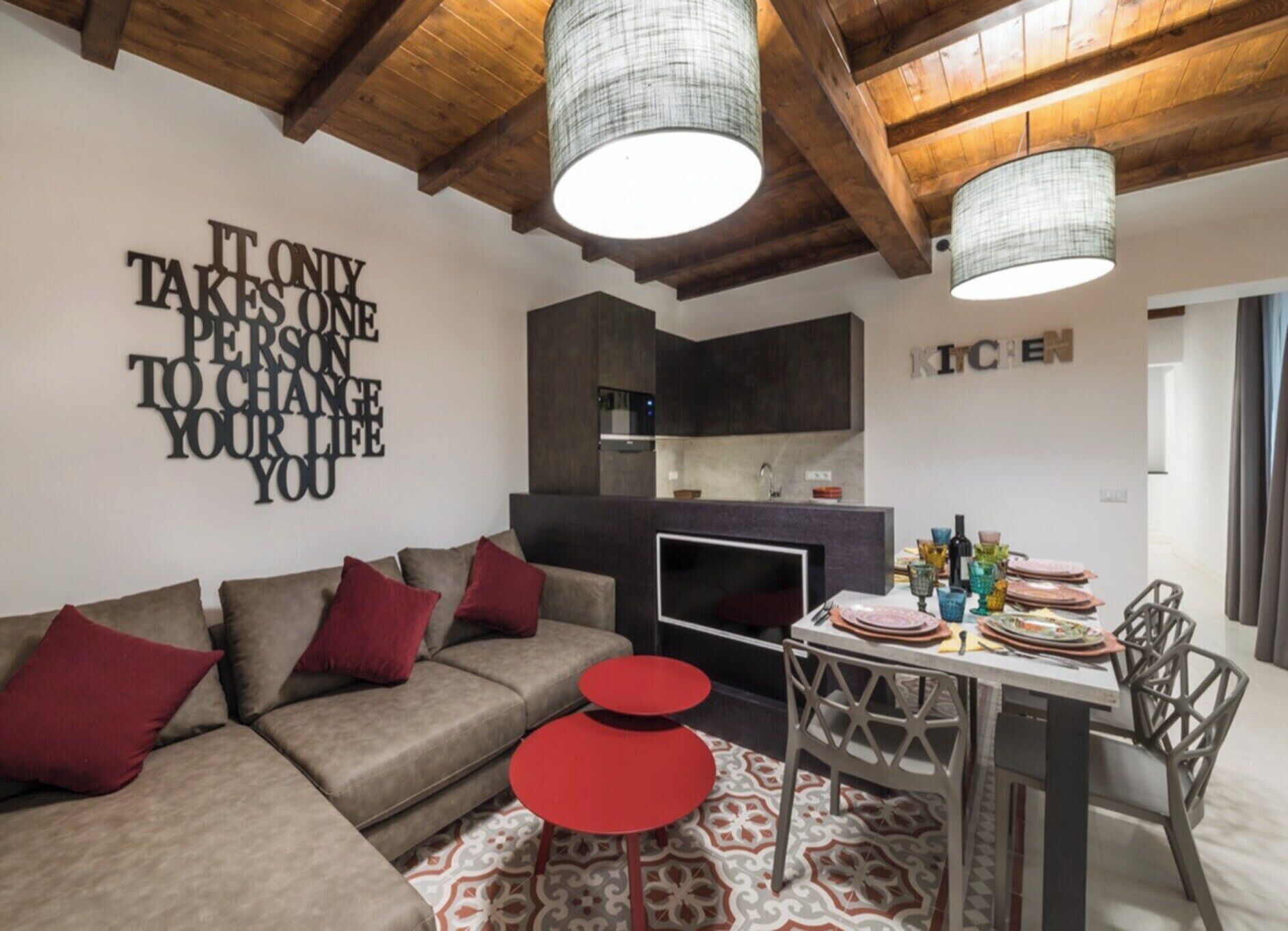 A modern living room with a sofa, red accents, a dining table with chairs, and a kitchen in the background.