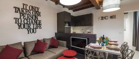 A modern living room with a sofa, red accents, a dining table with chairs, and a kitchen in the background.