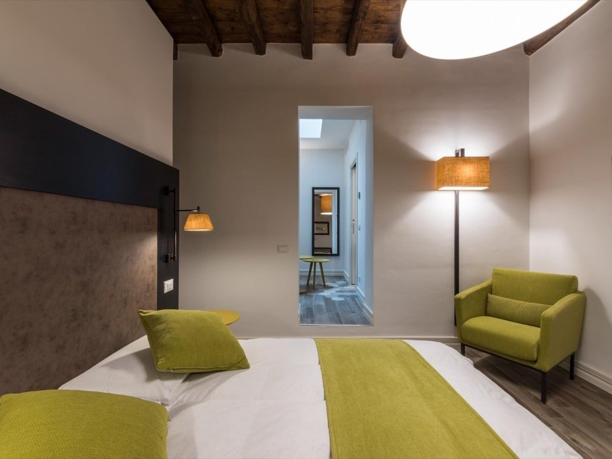 A hotel room with a bed, two green pillows, a green armchair, and a floor lamp.