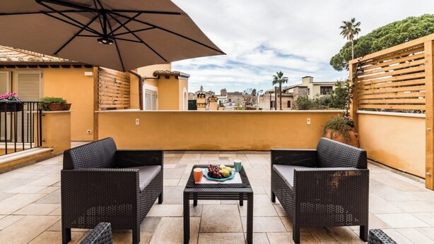 An outdoor patio with wicker furniture, a table with fruit, and a large umbrella. - Dolcevita Apartments (Roma)