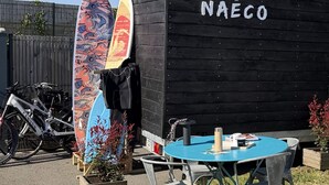 A black building with 'NAECO' written on it, a surfboard leaning against it, a bicycle, and a round table with chairs. - Naéco's Tiny Room (Plouharnel)
