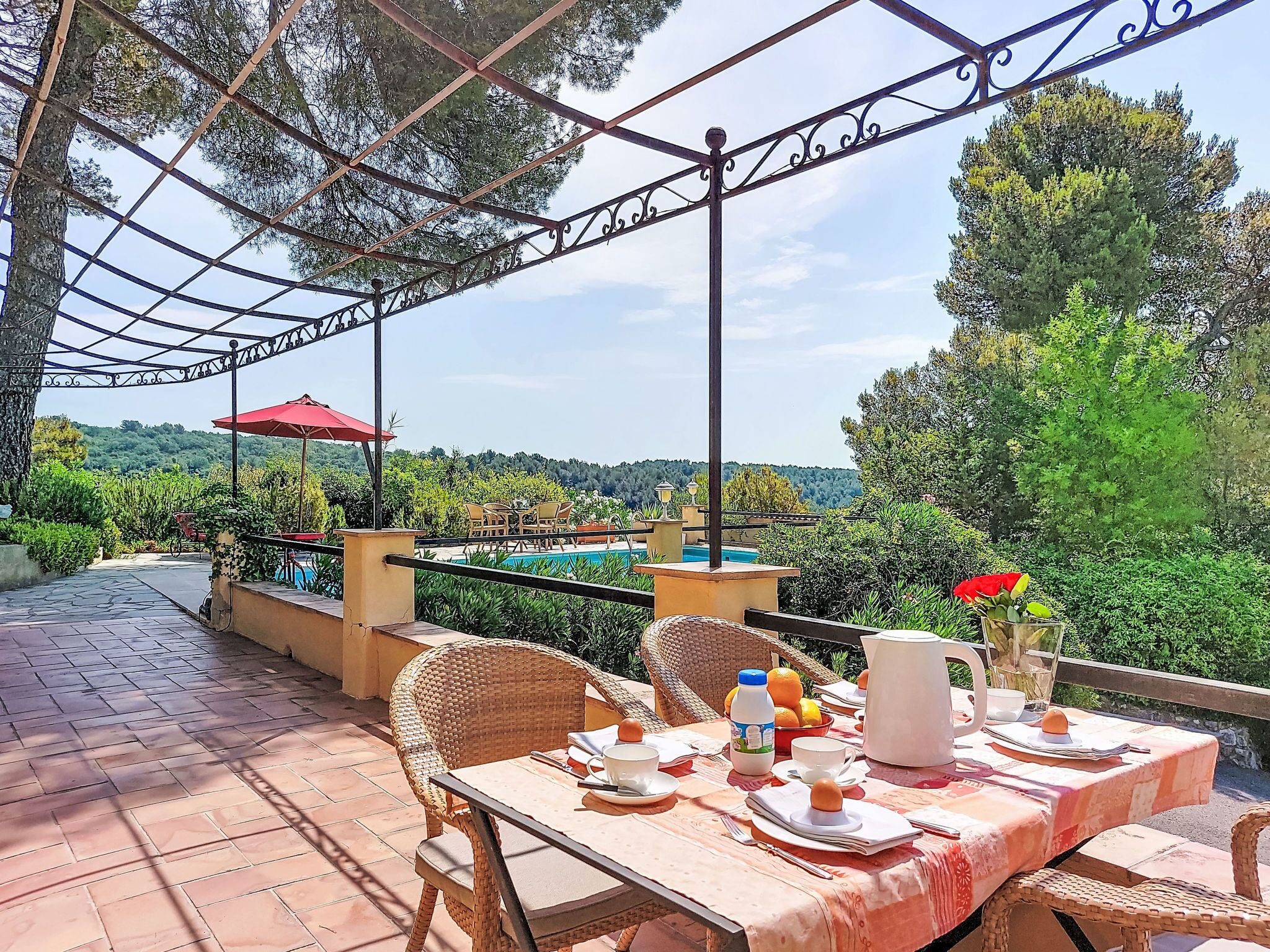 An outdoor dining area with wicker chairs, a table set for two, and a view of a pool and greenery.
