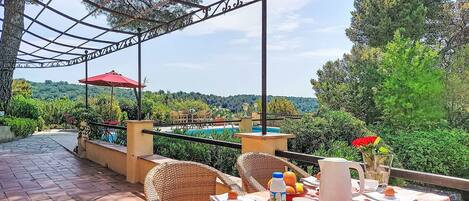 An outdoor dining area with wicker chairs, a table set for two, and a view of a pool and greenery.