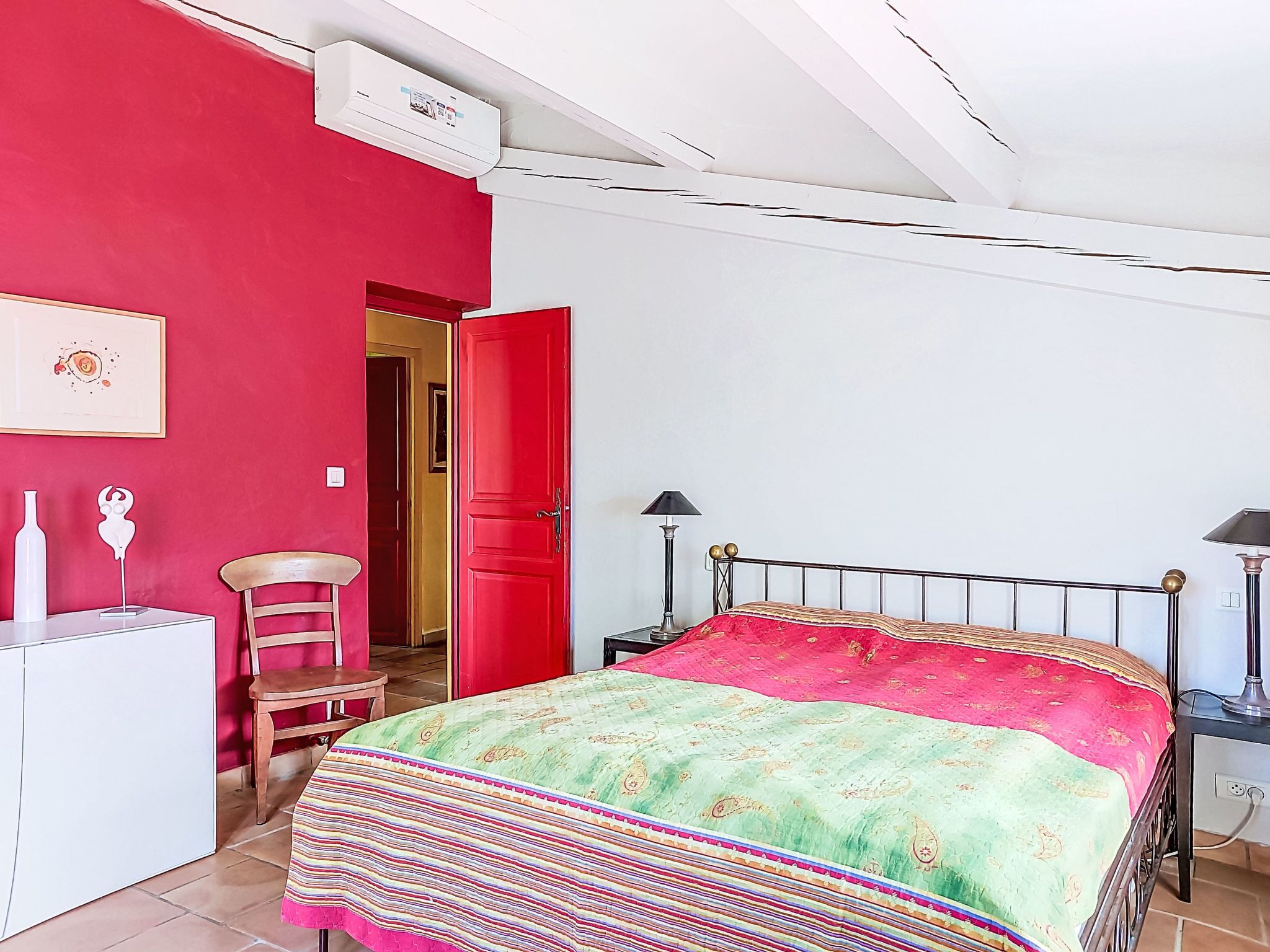 Um quarto com uma cama, uma cadeira, uma escrivaninha, um abajur e uma porta vermelha.