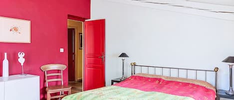 A bedroom with a bed, a chair, a desk, a lamp, and a red door.