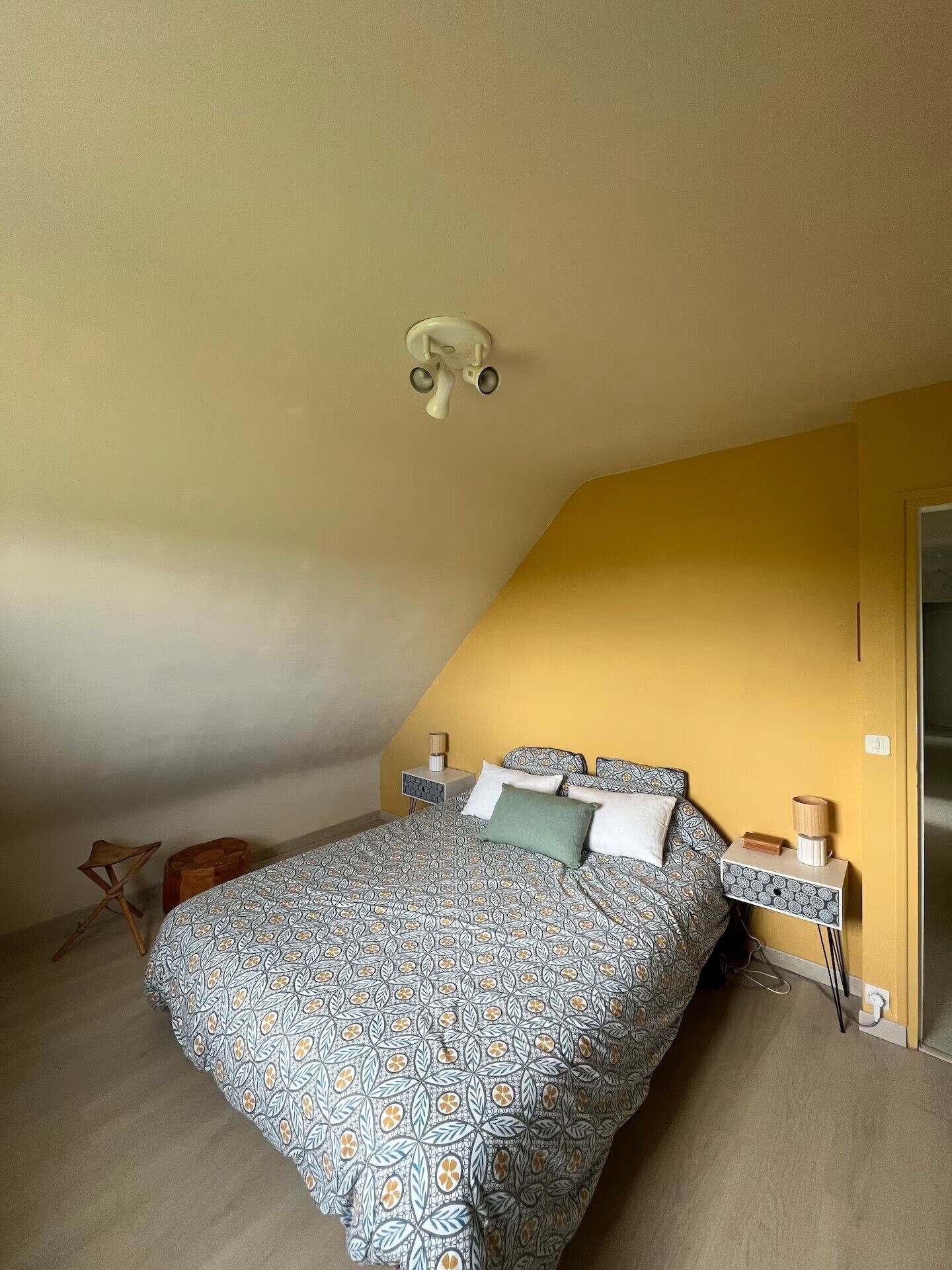 Ein Schlafzimmer mit schrägen Wänden, einem Bett mit gemusterten Bettbezügen, Nachttischen mit Lampen und einem hölzernen Hocker.