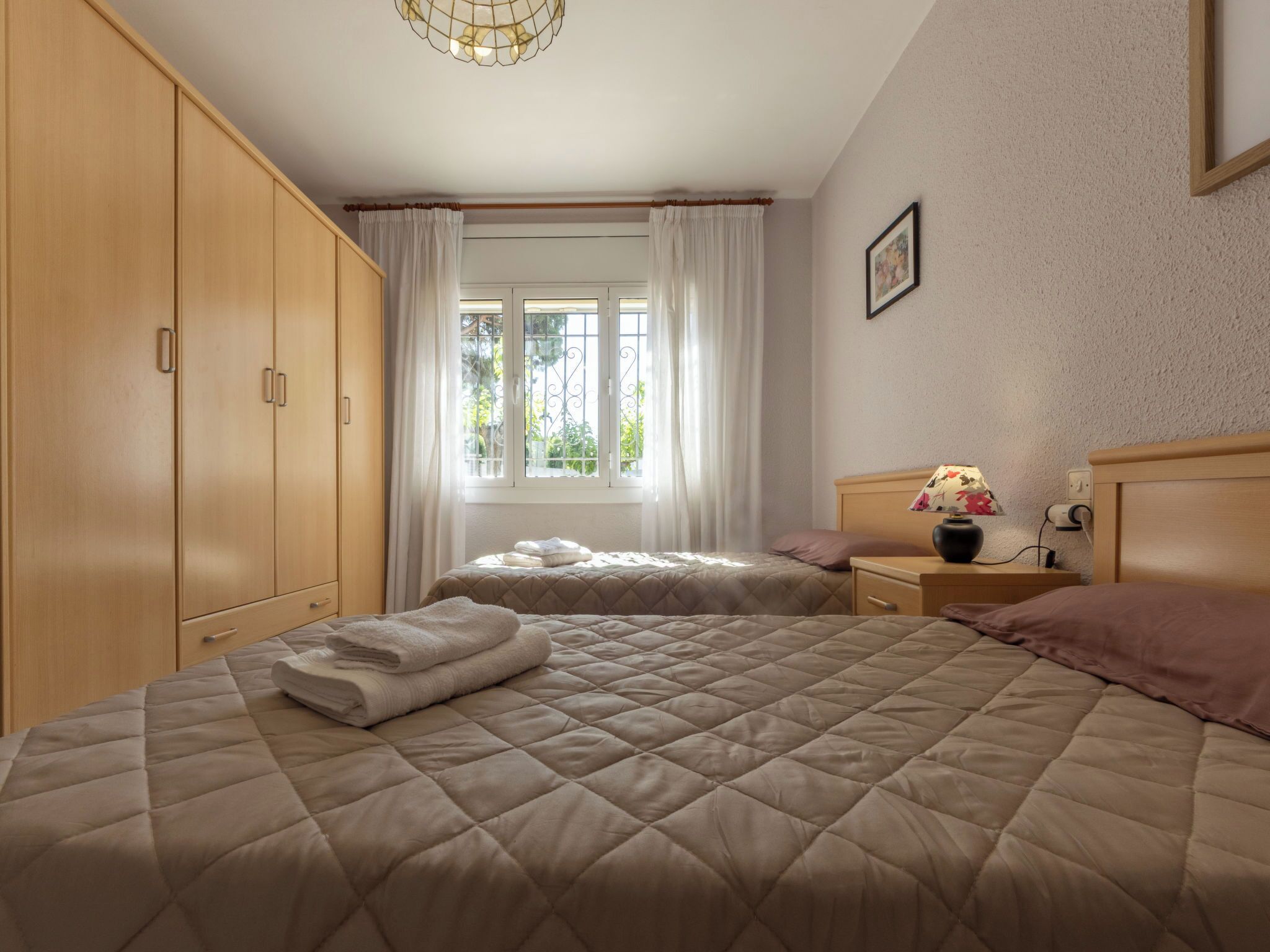 Une chambre avec deux lits, une armoire, une fenêtre avec des rideaux, une table de chevet avec une lampe et un tableau accroché au mur.