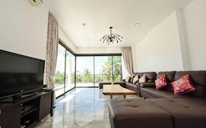 A modern living room with a flat-screen TV, a sofa set, a coffee table, and a large window offering a view of palm trees. - Skyline Seaview Villa-5 Bedrooms (จ.สุราษฎร์ธานี)
