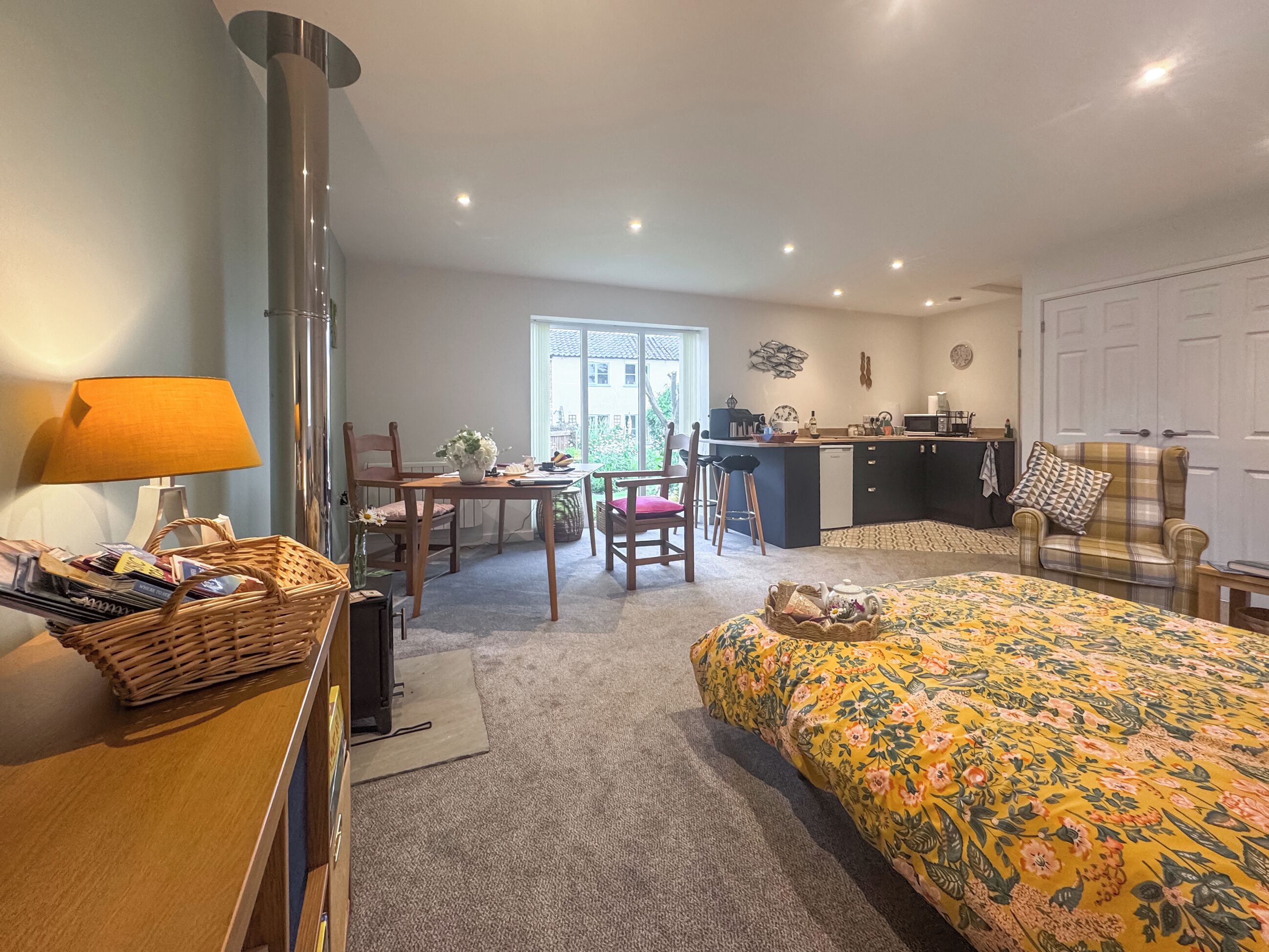 A cozy room with a bed, a desk, a chair, a lamp, a fireplace, and a kitchen area.