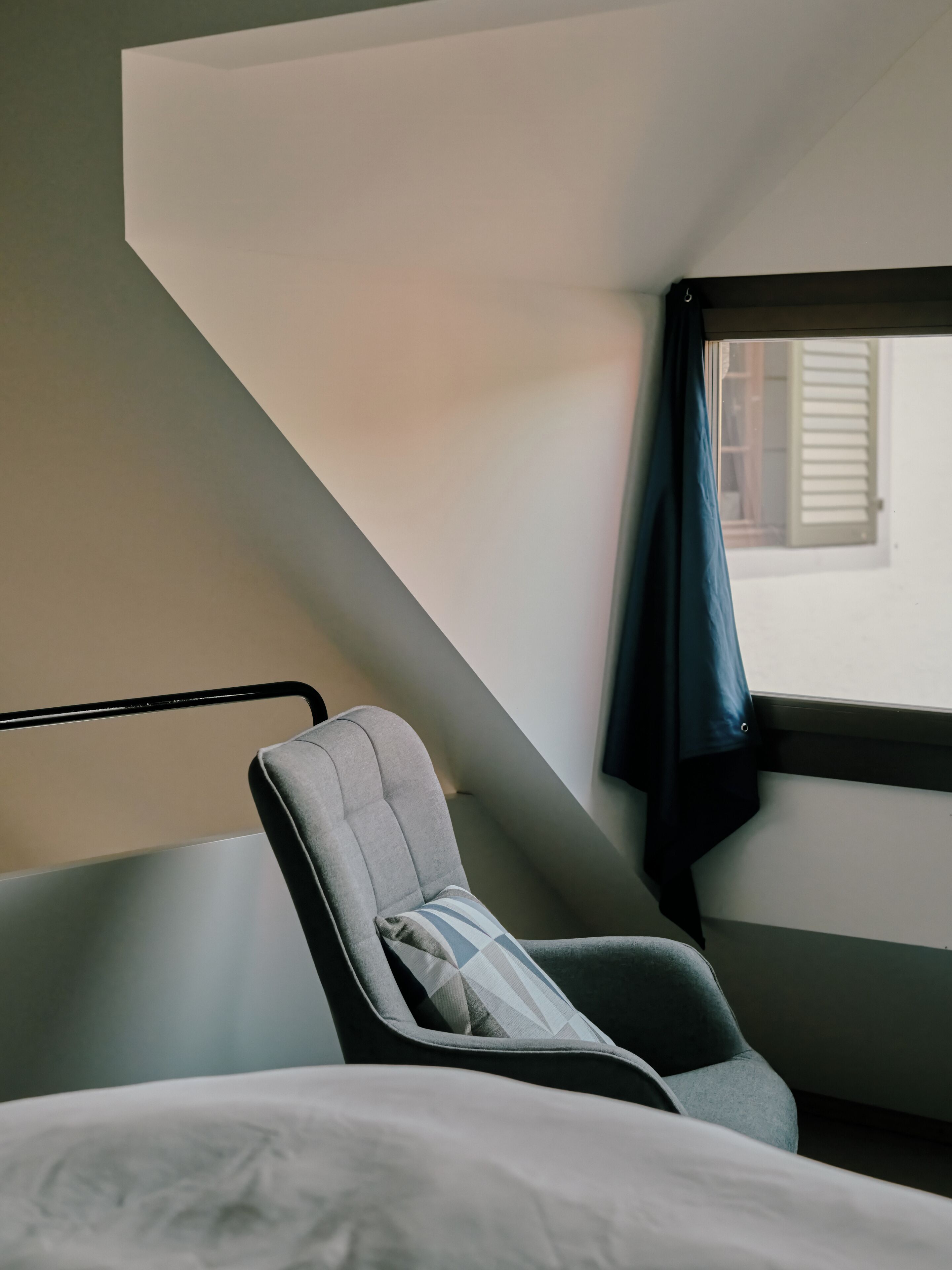 A hotel room with a bed, a chair with a pillow, a window with a curtain, and a view of the outside.