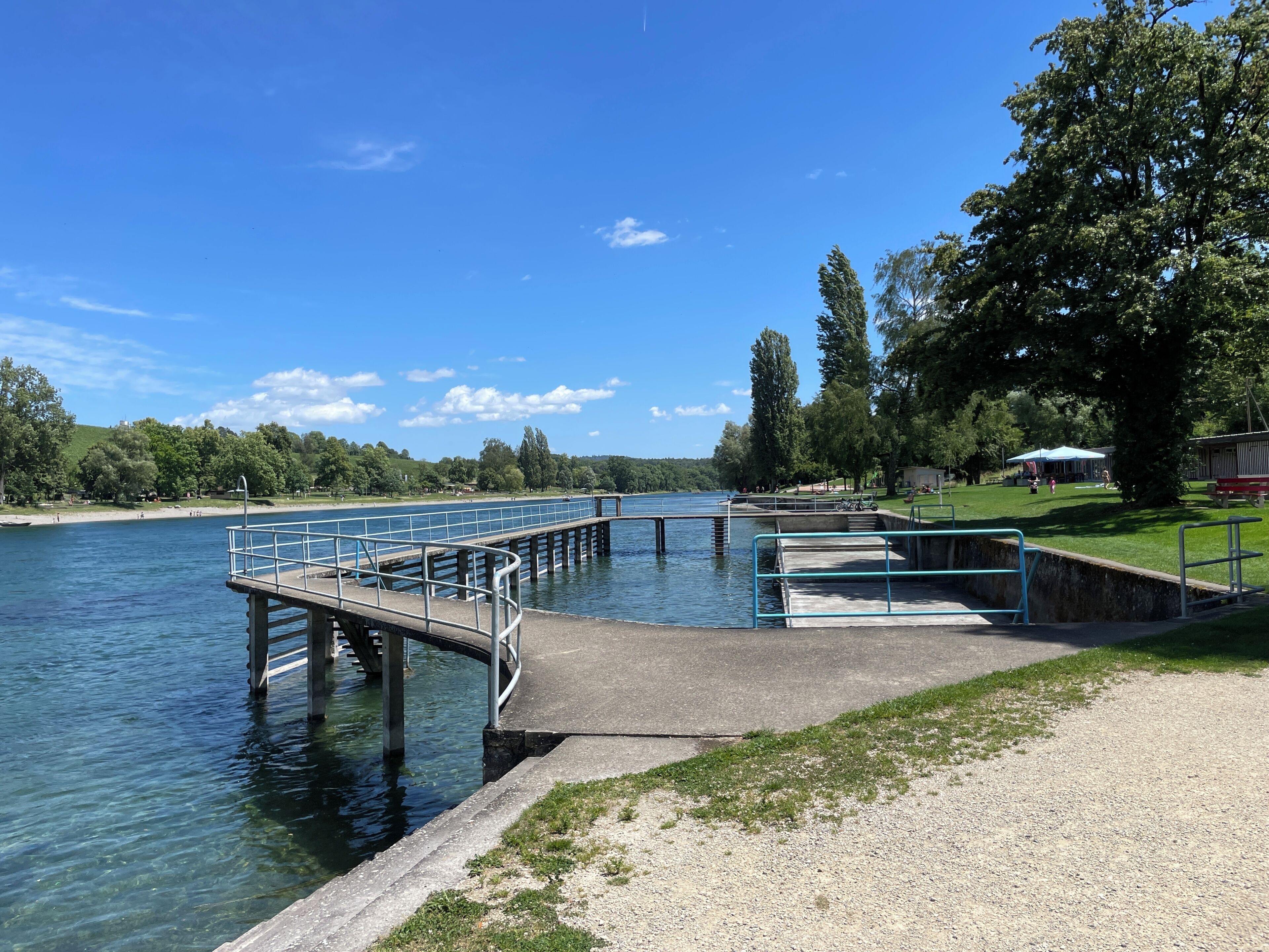Ein Park am Flussufer mit einem Betonweg, einer Metallgeländer und einer Rasenfläche.