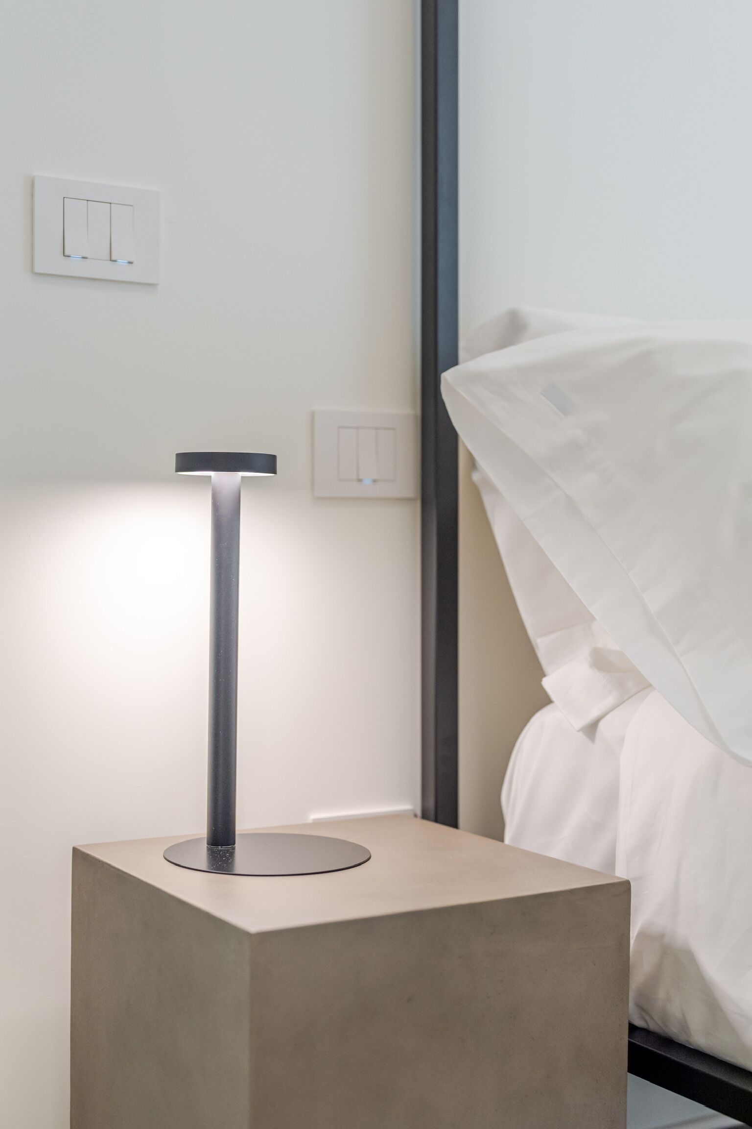 A modern bedside table with a floor lamp, a bed with white bedding, and a wall switch.