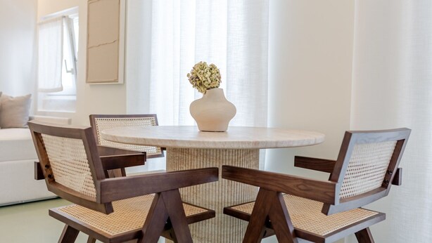 A dining area with a round table, four chairs, a vase with flowers, and a sofa in the background. - Garibaldi Luxury Suites - Graecalis (Noci)
