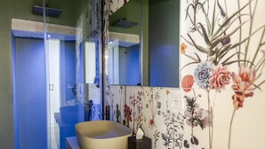 A bathroom with a large mirror, a beige sink, a tissue box, and a floral wall decoration. - Garibaldi Luxury Suites - Graecalis (Noci)