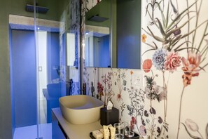 Ein Badezimmer mit einem großen Spiegel, einem beige farbenen Waschbecken, einer Papiertuchbox und einer Wanddekoration mit Blumenmuster.