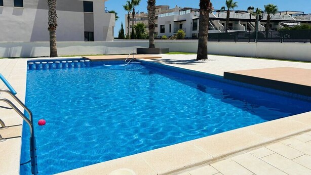 A rectangular swimming pool with clear blue water, surrounded by a tiled deck and palm trees. - Stunning 3-bedroom apartment with a pool – Ideally located in La Zenia! (Orihuela)