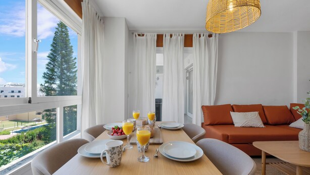 Ett modernt matområde med ett bord dukat för frukost, en soffa och utsikt över ett träd och några byggnader.