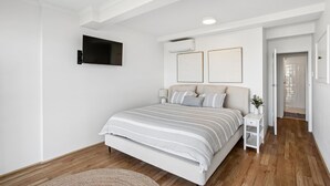 Un dormitorio con una cama, un televisor montado en la pared, una mesita de noche y una puerta que da a otra habitación.