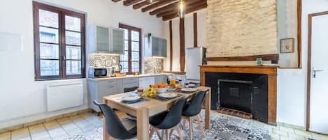 A kitchen with a stone wall, wooden beams, a dining table with chairs, and a fireplace.