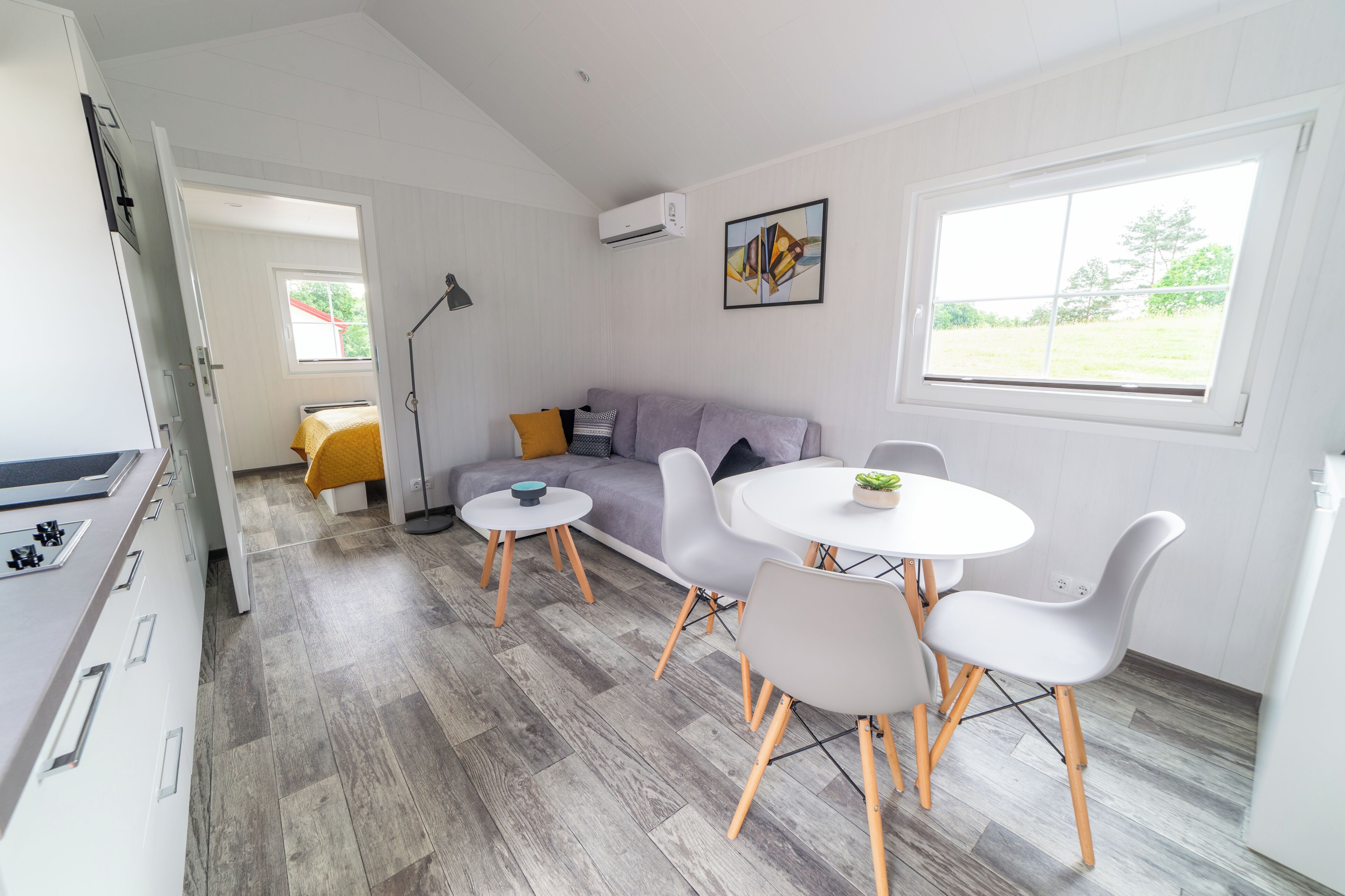 Ett modernt kök med en inbyggd spis, en matplats med ett runt bord och stolar, en grå soffa med kuddar och ett stort fönster med utsikt över omgivningen.