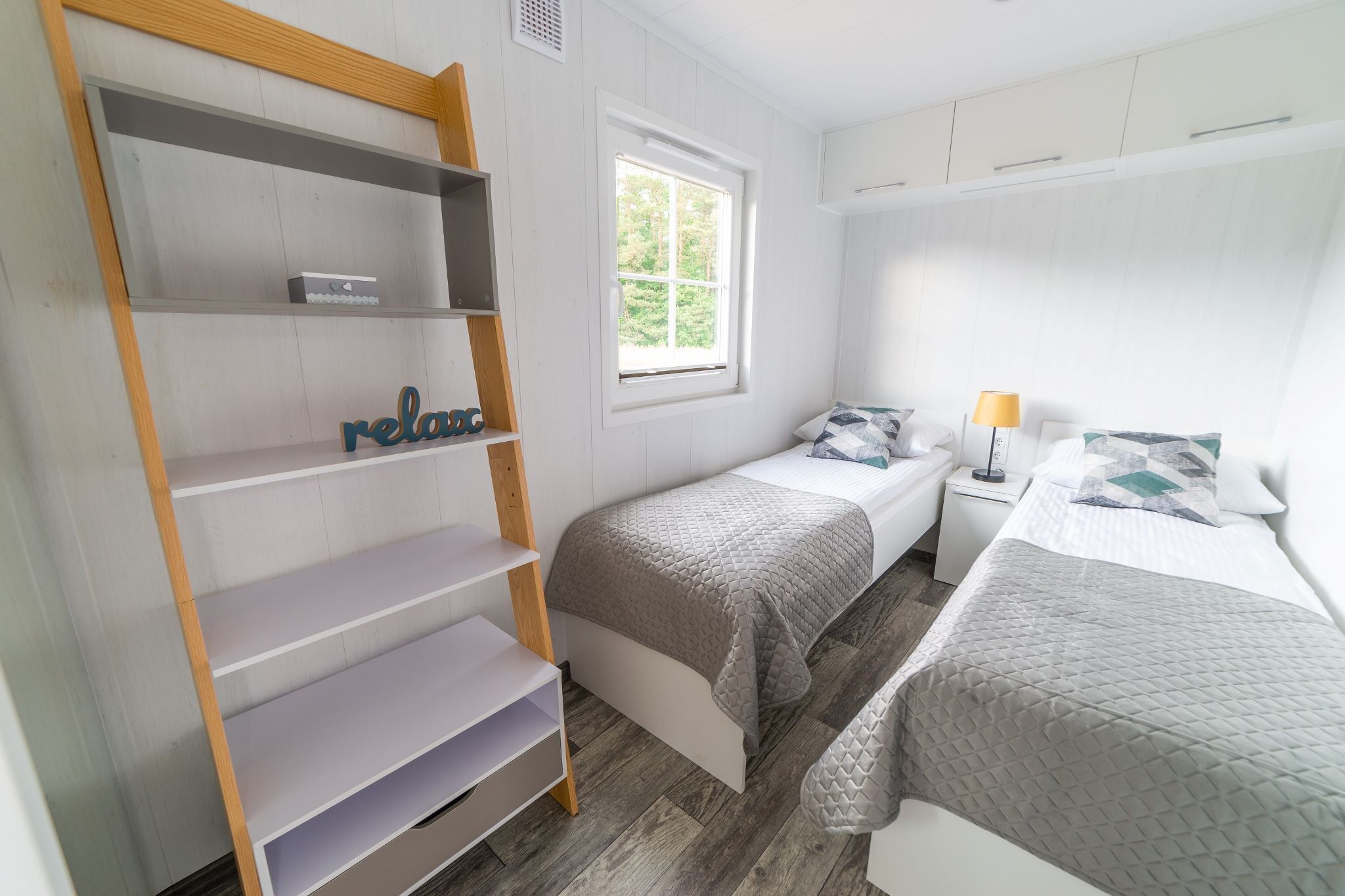 Un dormitorio con cama, mesita de noche, lámpara, ventana y estantería.