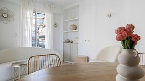 Moderni olohuone, jossa on sohva, ruokapöytä, maljakko kukkineen ja ikkunasta avautuva näkymä parvekkeille.