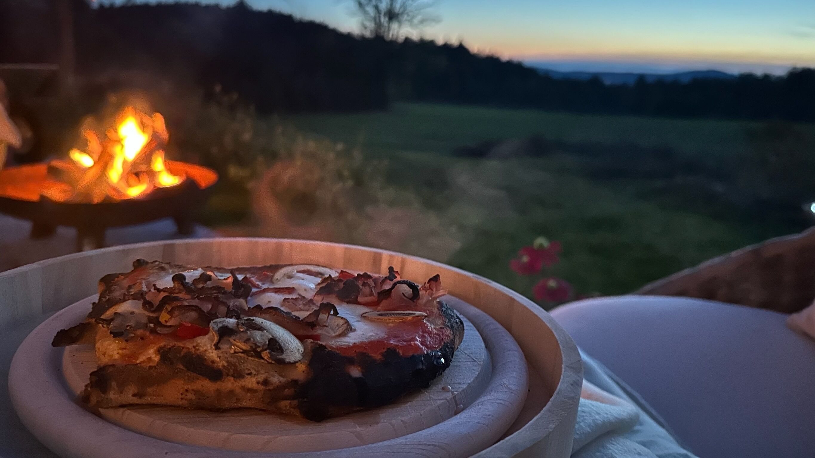 Una porción de pizza sobre un plato de madera, con fuego al fondo.
