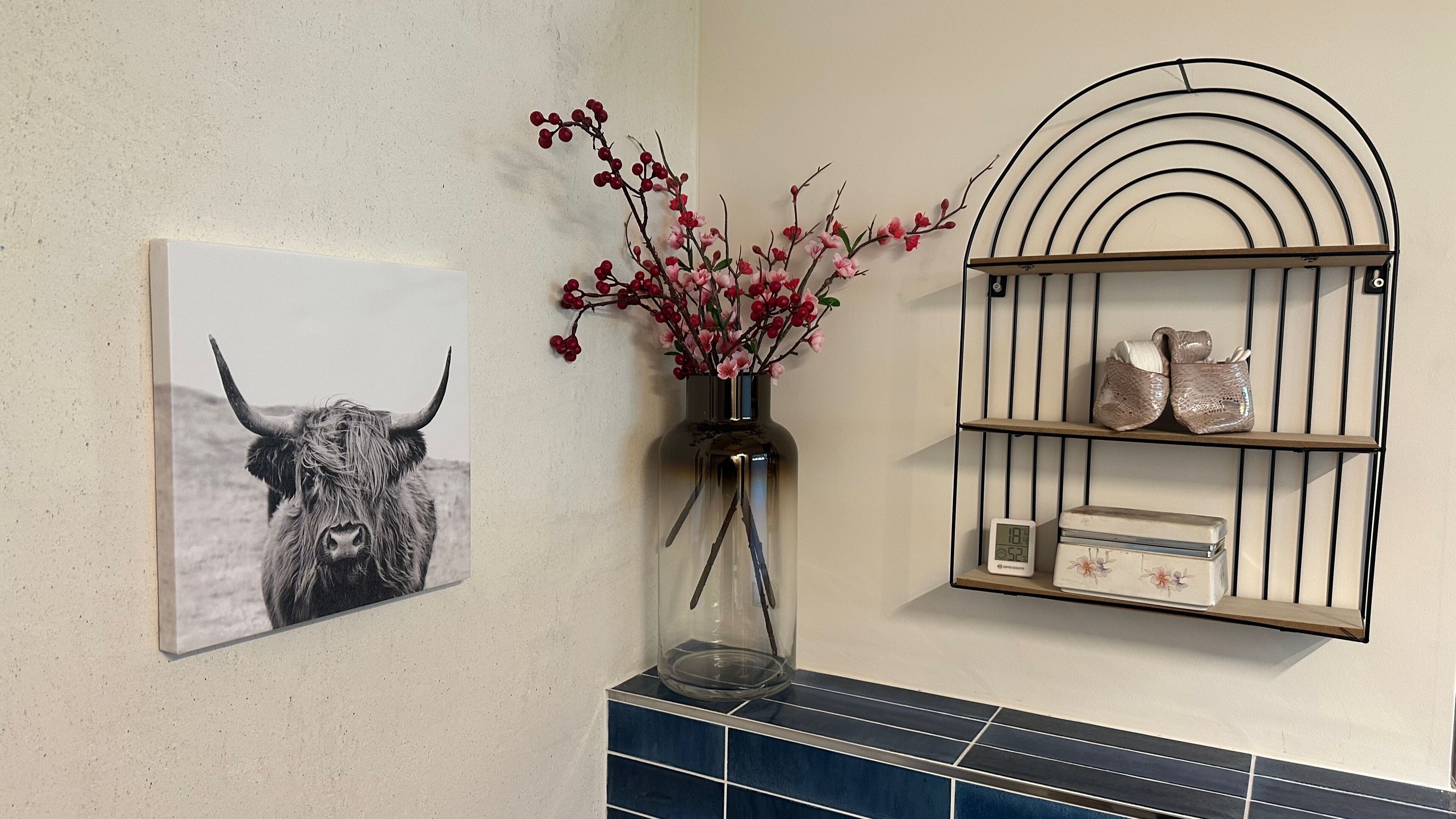 Une salle de bain avec une étagère fixée au mur où sont rangés les articles de toilette, un tableau représentant un taureau et un vase avec des fleurs roses.