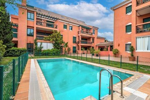 A swimming pool with a metal ladder, surrounded by a green fence, and adjacent to a multi-story building with balconies. - LXR Marbesa 1-Bedroom Near the Sea (Marbella)