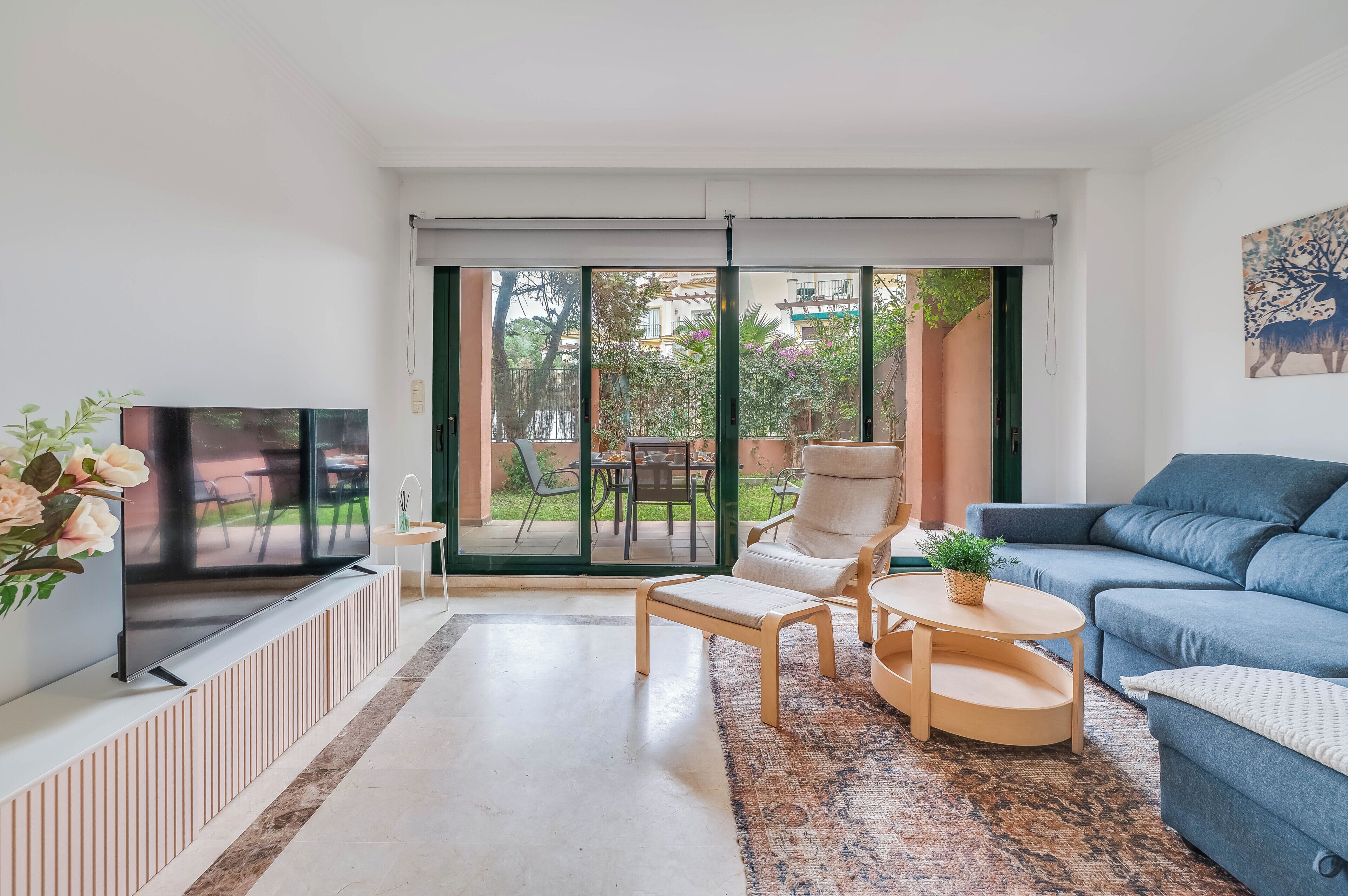 A modern living room with a flat-screen TV, a blue sofa, a white armchair, and a small round table with a plant.