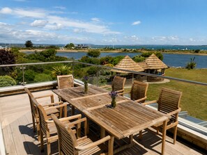 A wooden deck with a table and chairs, overlooking a body of water and a grassy area with thatched-roof structures. - Fantastic Large Chichester Harbour View House. 2 minutes to beach. (Hayling Island)