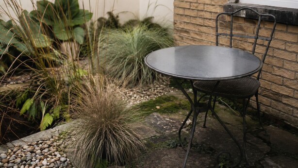 Une table ronde en métal, accompagnée d’une seule chaise, est placée à l’extérieur, avec un mur de briques et une fenêtre en arrière-plan.