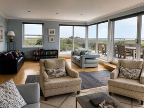 A spacious living room with a large window, a bookshelf, and a balcony with outdoor furniture. - Fantastic Large Chichester Harbour View House. 2 minutes to beach. (Hayling Island)