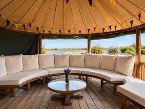 A covered outdoor seating area with a curved sofa and a round table, overlooking a natural landscape. - Fantastic Large Chichester Harbour View House. 2 minutes to beach. (Hayling Island)