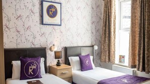 A hotel room with two beds, each with a purple bedspread and matching pillow, a nightstand with a lamp, a framed logo on the wall, and a window with curtains. - Standard Twin Room (Norwich)