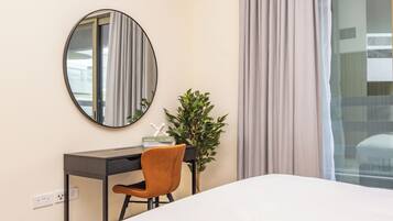 A hotel room with a bed, a desk, a chair, a mirror, a plant, and a window with curtains.
