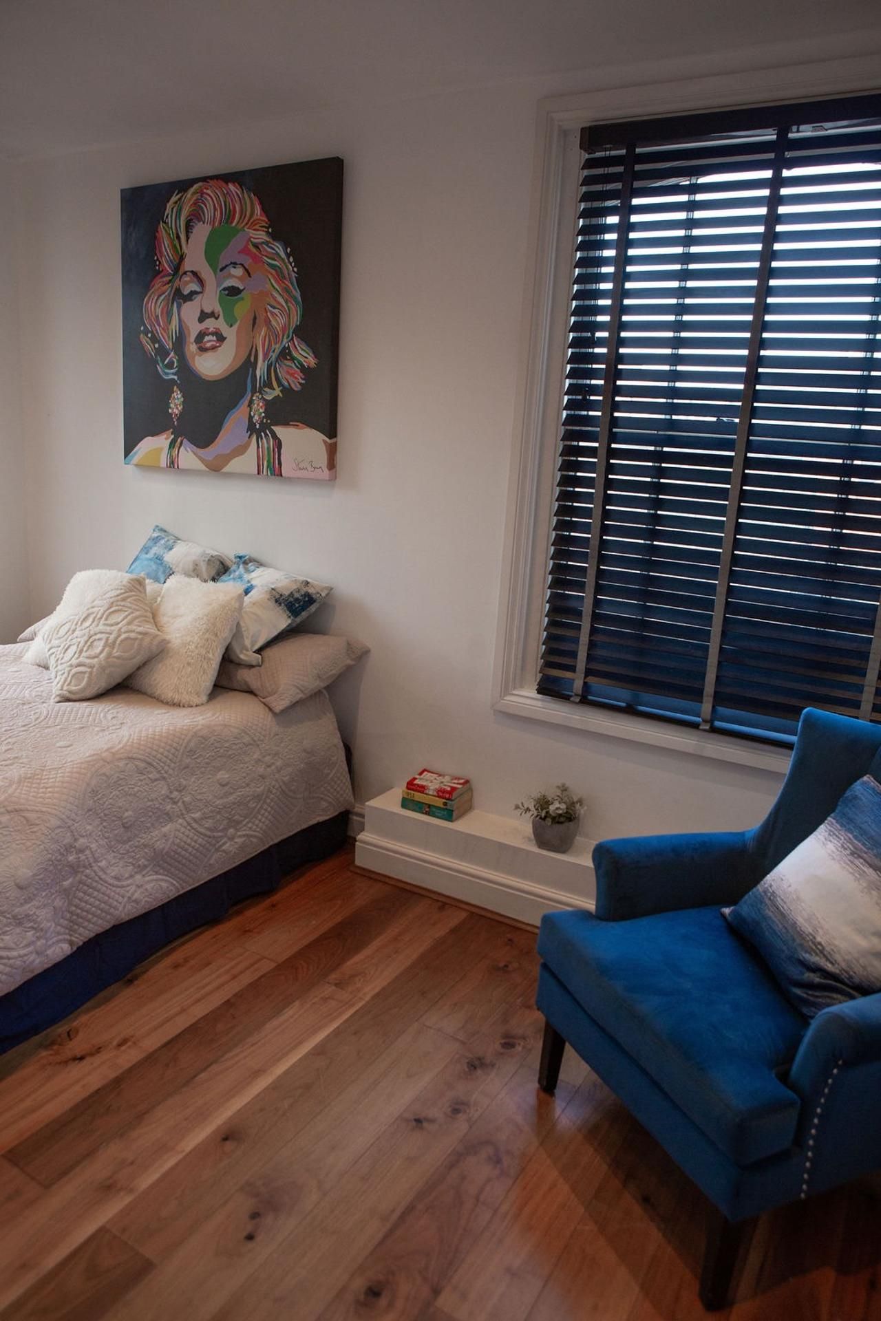 A bedroom with a bed, a blue armchair, a window with blinds, and a painting on the wall.