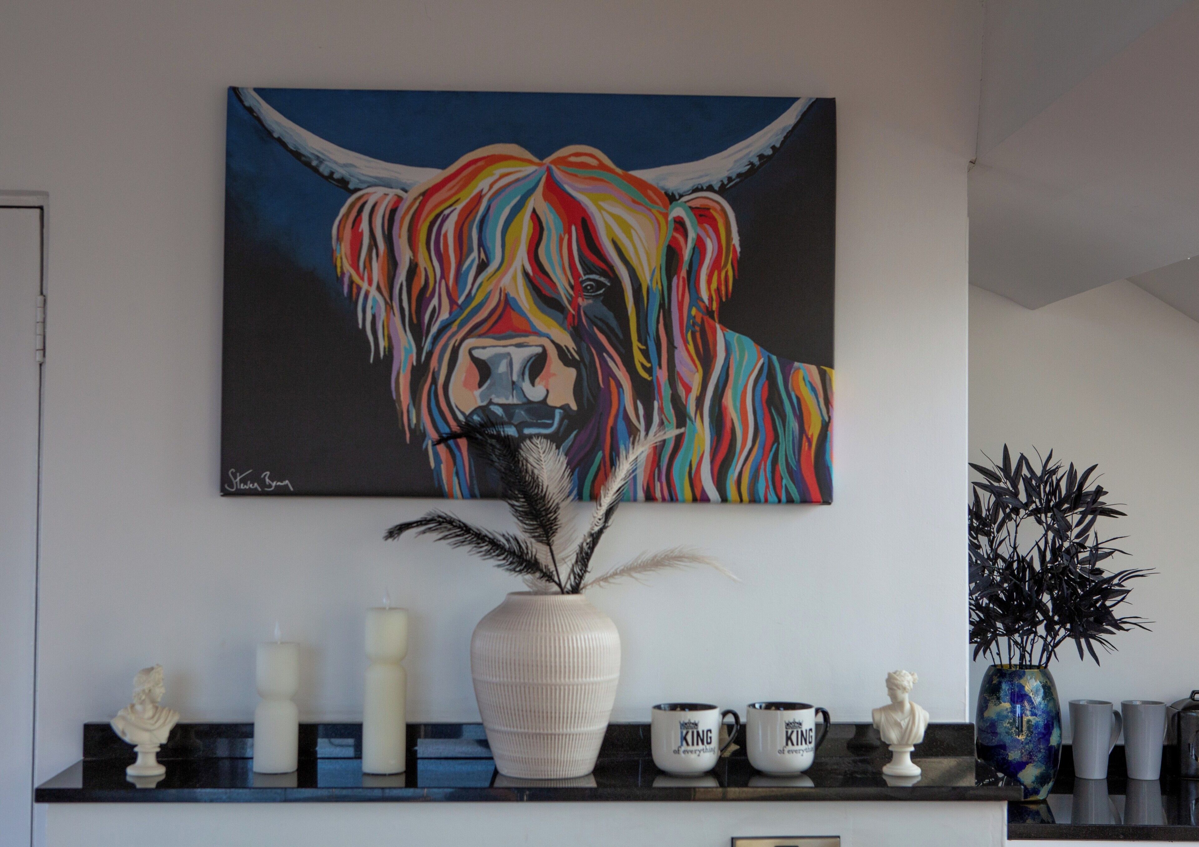 Pintura de um touro colorido numa parede, um vaso branco com penas e uma bancada preta com diversos objetos.