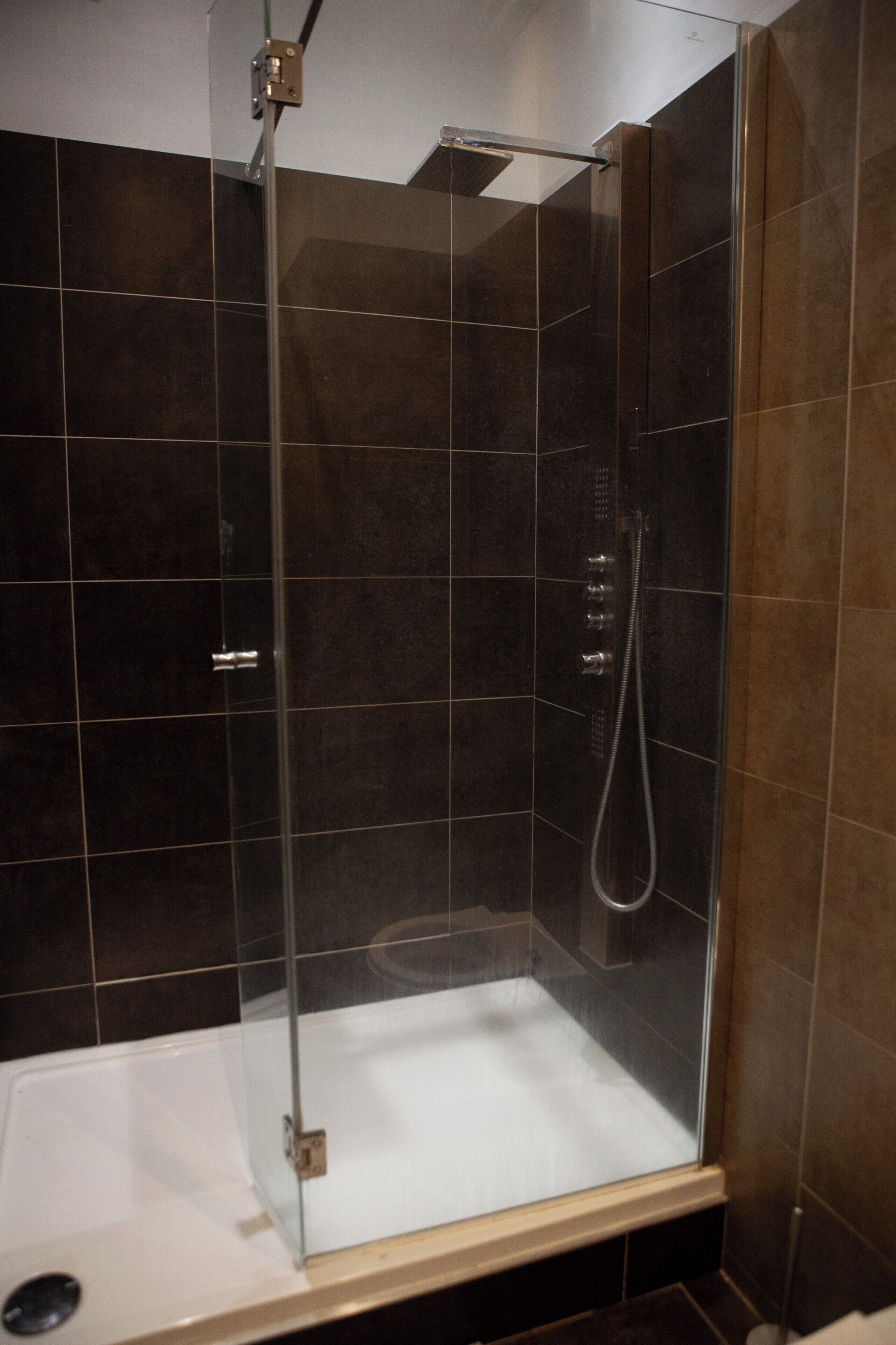 Cabina de ducha con paredes de vidrio, una bañera blanca, paredes de baldosas oscuras y un panel de control de la ducha a la vista.