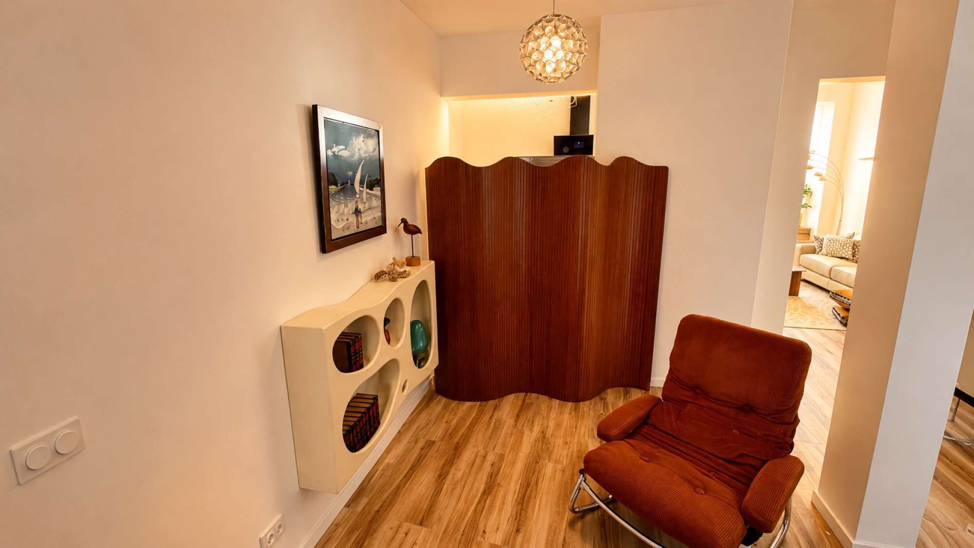 Ein Zimmer mit Holzboden, einem roten Sessel, einem weißen Konsolentisch mit Ablagen, einem gerahmten Bild an der Wand und einer Holzvertäfelung.